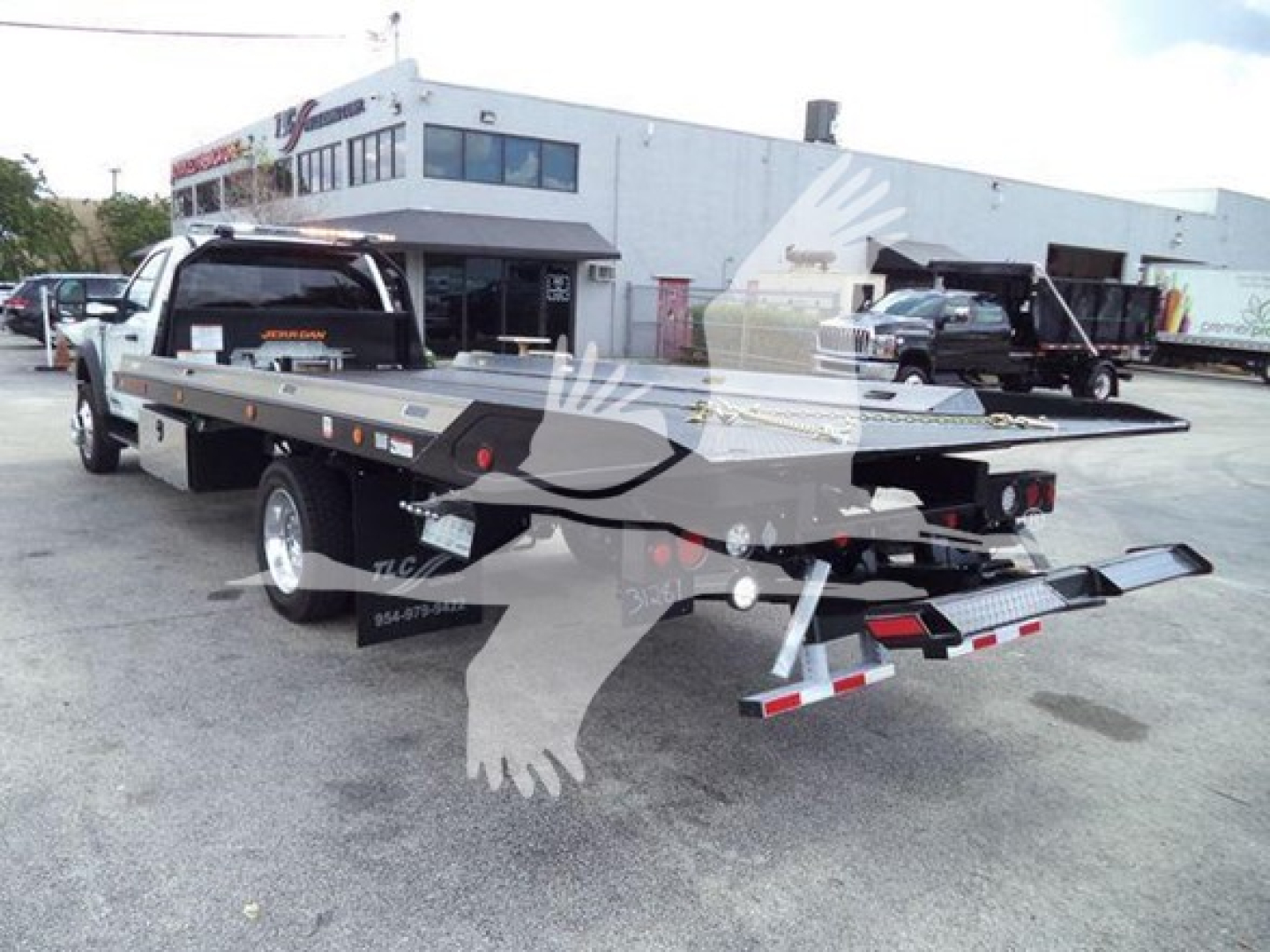 Flatbed tow truck with a long metal deck, rear lighting, and hydraulic tilt mechanism visible; dealership building and other trucks in background.