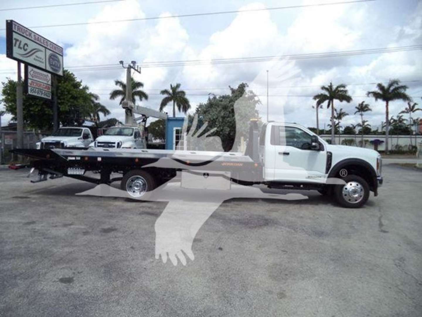 Ford F-550 rollback tow truck in a parking lot, white cab and long wheelbase, flatbed with winch mounts, palm trees in background