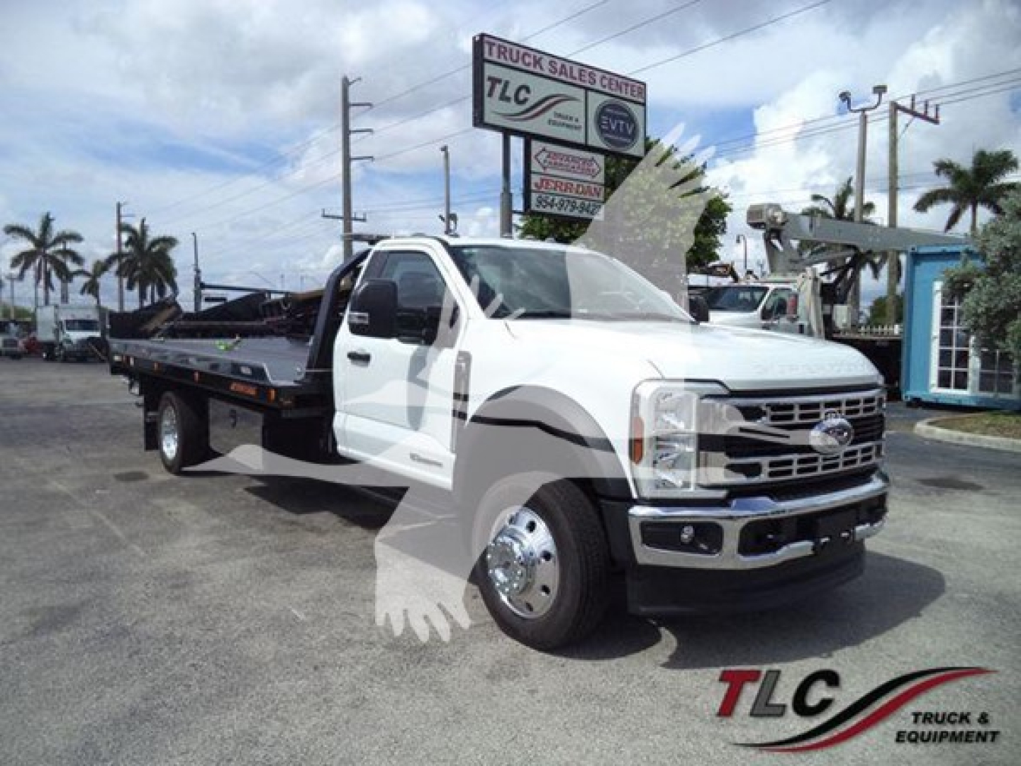 Truck image showing a white 2024 Ford F550 Rollback tow truck with a flatbed, front grille, chrome bumper, and black flatbed; dealership signage in background.