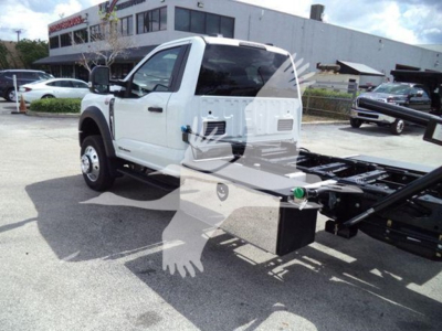 Front-left view of a white 2024 Ford F550 rollback truck with a crane/chassis frame in the bed area. Visible text on site signage in background.