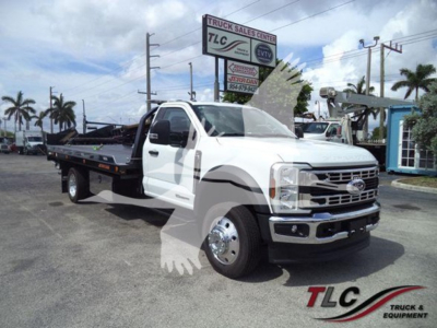 Truck image showing a white 2024 Ford F550 Rollback tow truck with a flatbed, front grille, chrome bumper, and black flatbed; dealership signage in background.