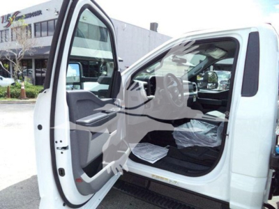 Truck cab interior visible through open driver door of a white Ford F-550 rollback truck, steering wheel and dashboard seen, papers on floor mat, building in background.