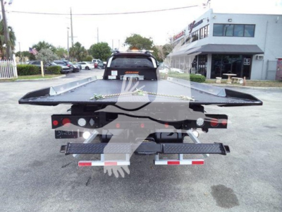 Ford F-550 rollback tow truck rear view showing flatbed with side rails, lights, and bumper, dealership in background.