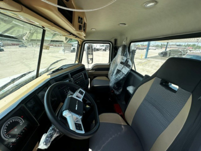 Cab interior of a 2027 Mack GR64F showing steering wheel with control stalks, dashboard, and two-tone cloth seats; plastic covering on the driver seat.