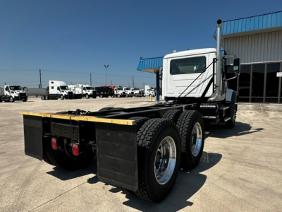 Truck chassis showing dual rear axles with fenders, fifth-wheel area, and cab in distance; white cab, black skirts, blue canopy over a dealership lot.