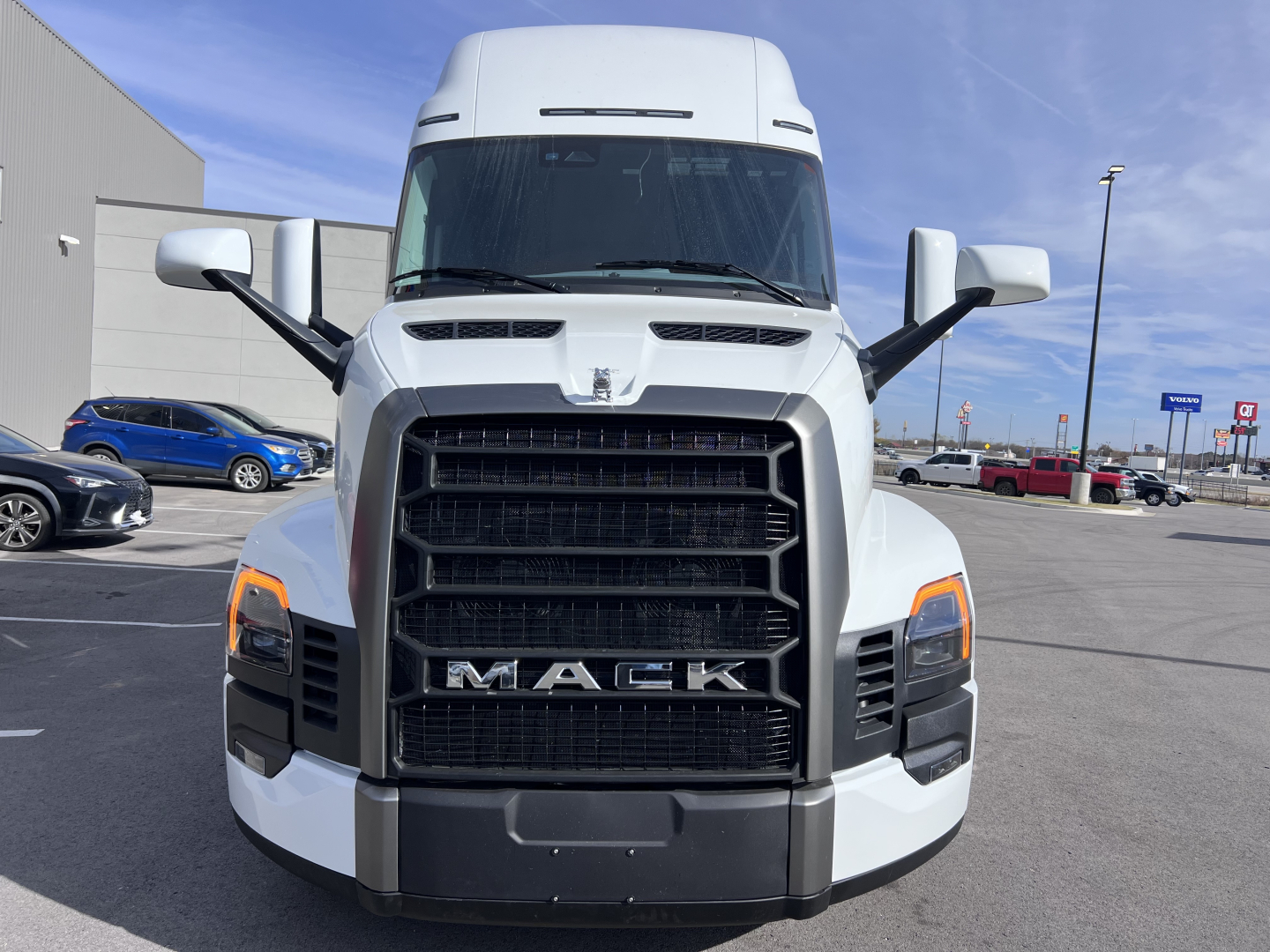 Front view of a white 2026 Mack PR64T76H truck with large black grille, prominent Mack logo, and side mirrors; showroom lot in background.