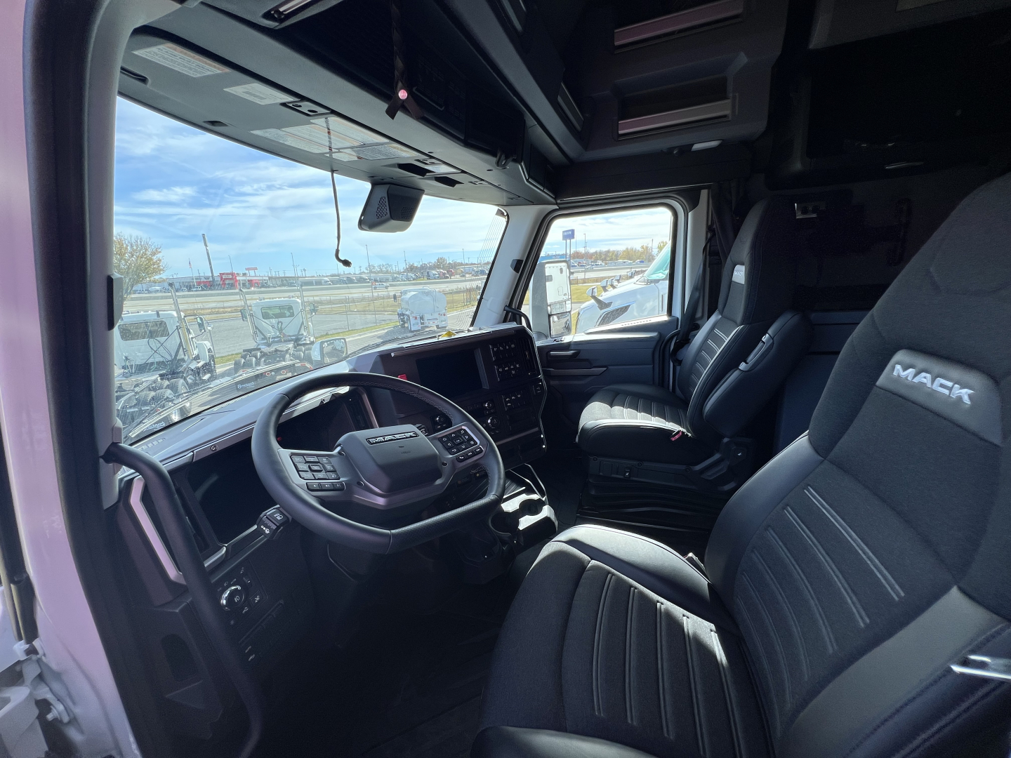 Truck cab interior view of a 2026 Mack PR64T76H showing steering wheel, large touchscreen display, driver and passenger seats, and dash controls.