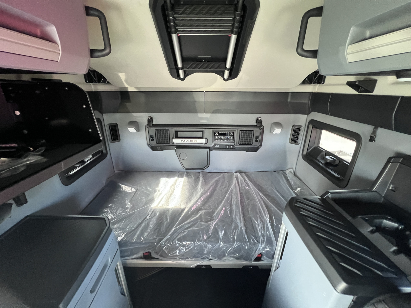 Cab interior of a new Mack PR64T76H tractor showing sleeper area with overhead cabinets and control panels, plastic-covered mattress, side windows, and overhead storage.