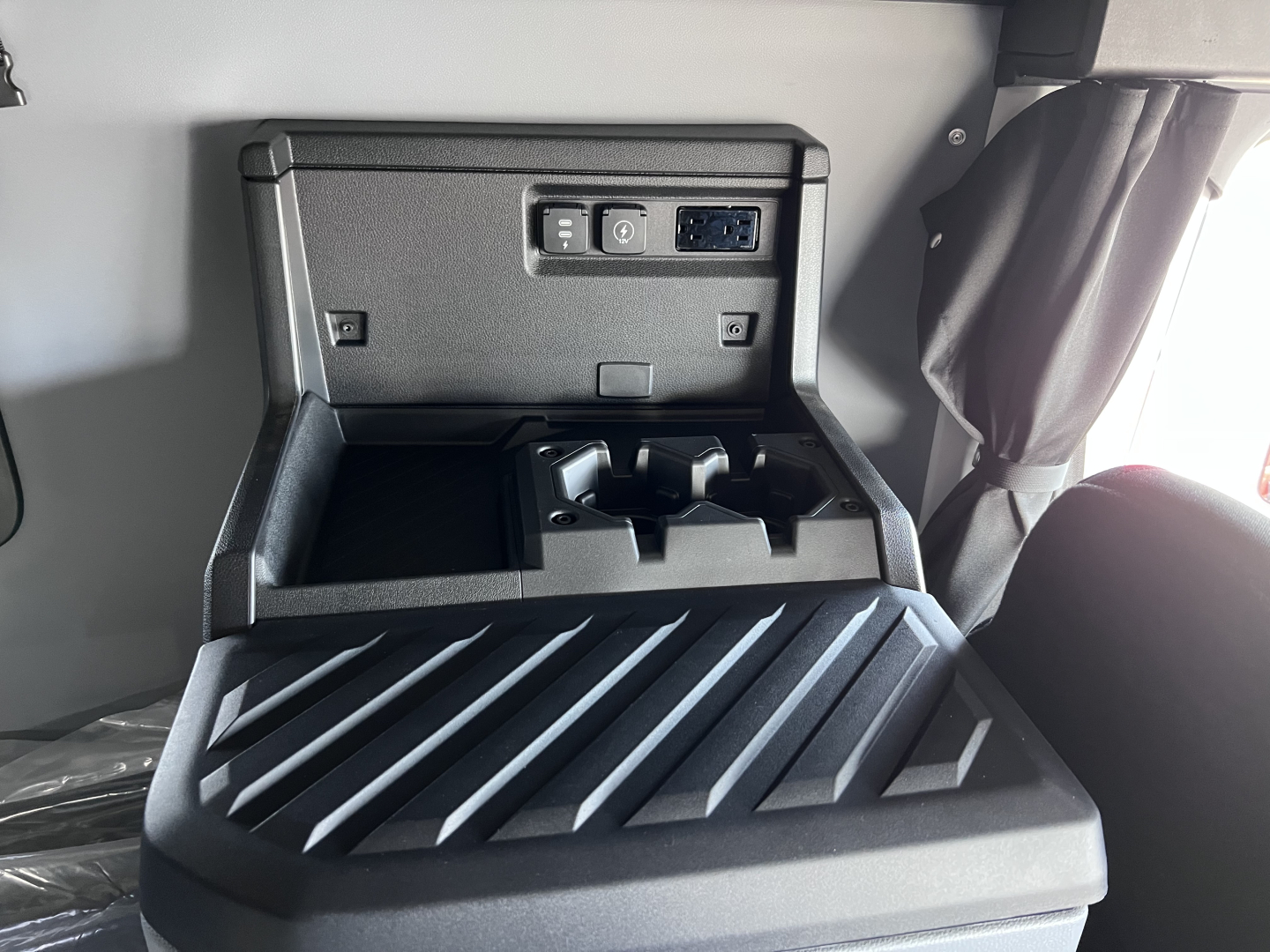 Cab interior storage area with power outlet panel and USB/12V ports in a Mack PR64T76H truck cabin, showing open console with organizer tray.