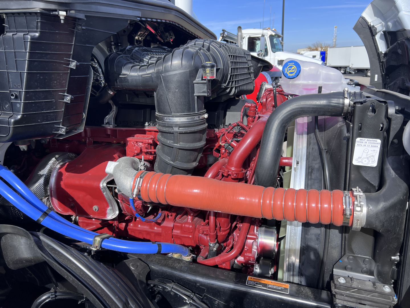 Engine bay of a 2026 Mack PR64T76H with red engine, orange flexible hose, black intake, blue hose, and radiator area visible.