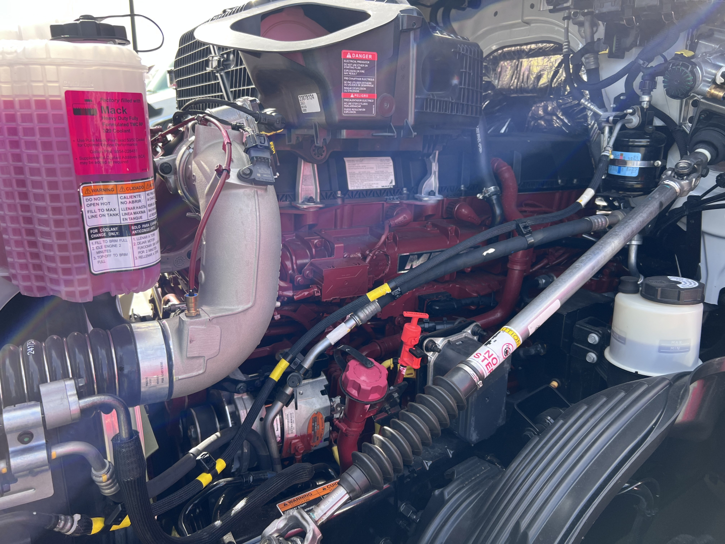 Engine bay of a 2026 Mack PR64T76H showing coolant reservoir, air intake, hoses, and various hydraulic lines. Readable labels on pink coolant reservoir and nearby components.