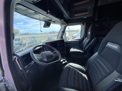 Truck cab interior view of a 2026 Mack PR64T76H showing steering wheel, large touchscreen display, driver and passenger seats, and dash controls.