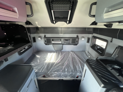 Cab interior of a new Mack PR64T76H tractor showing sleeper area with overhead cabinets and control panels, plastic-covered mattress, side windows, and overhead storage.