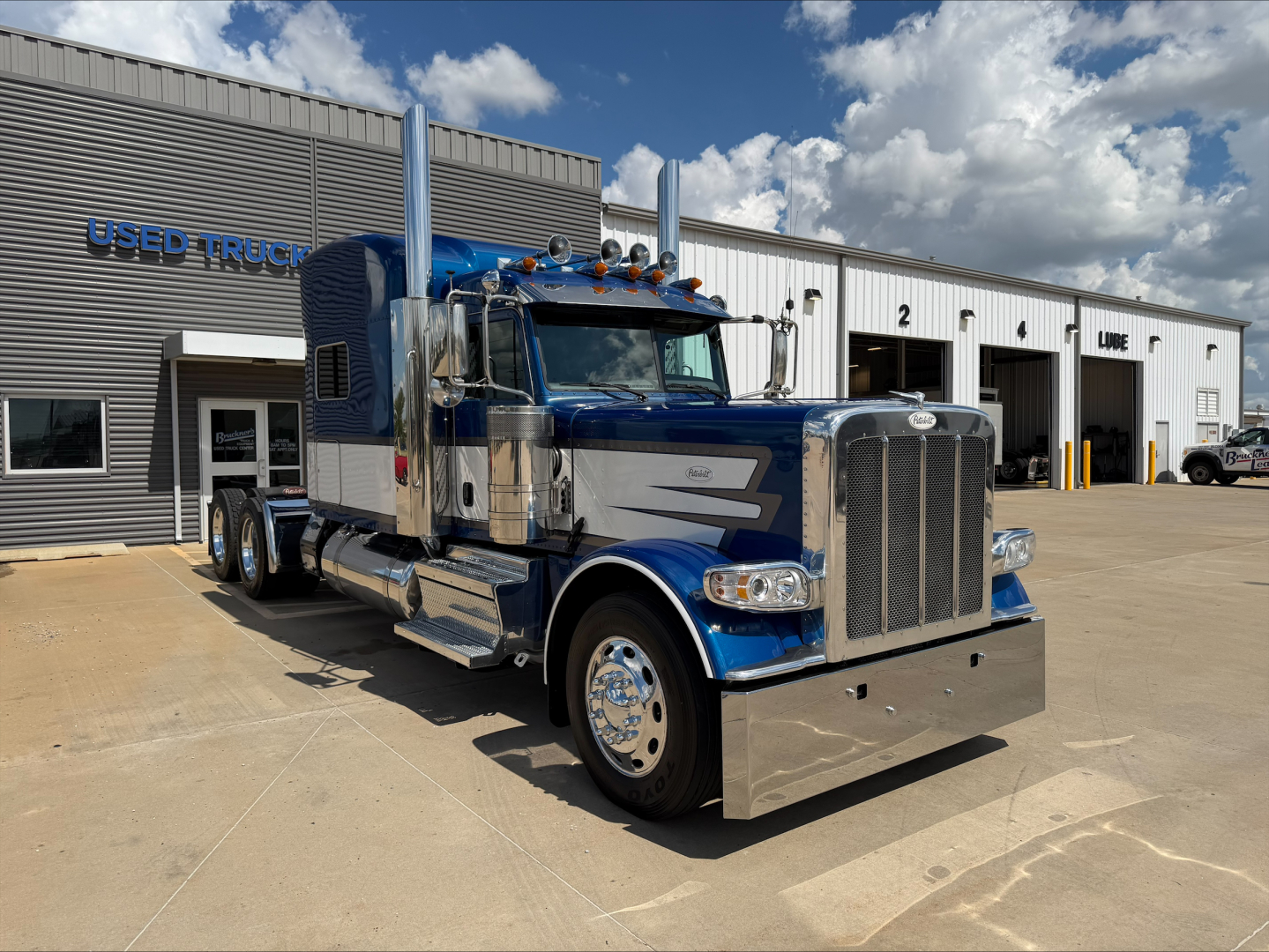 Peterbilt 389 conventional sleeper truck in blue and chrome, front three-quarter view, parked outside dealership.