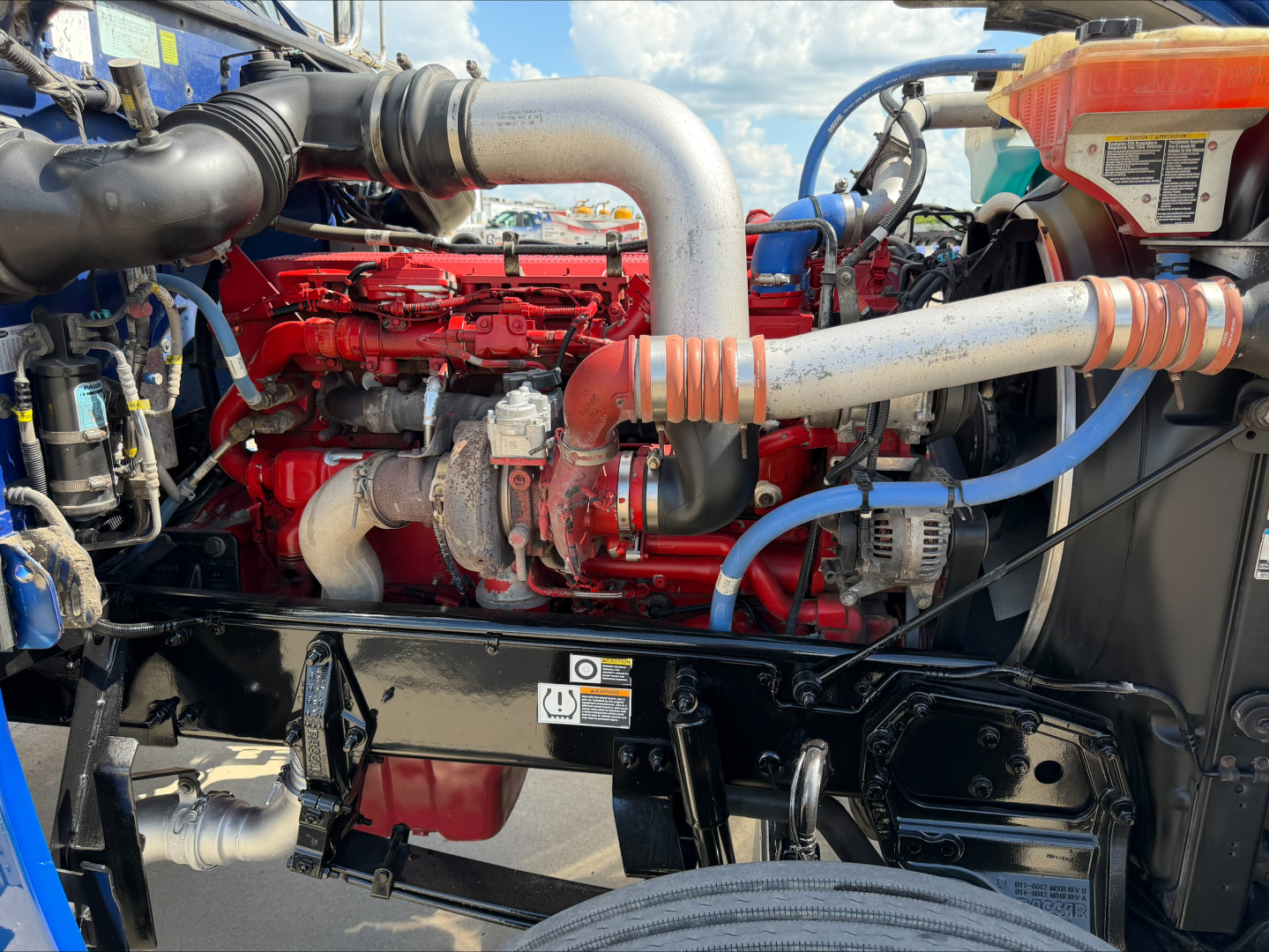 Peterbilt 389 engine bay showing large aluminum intake pipe, red engine block with hoses and electrical wiring, and blue coolant lines.