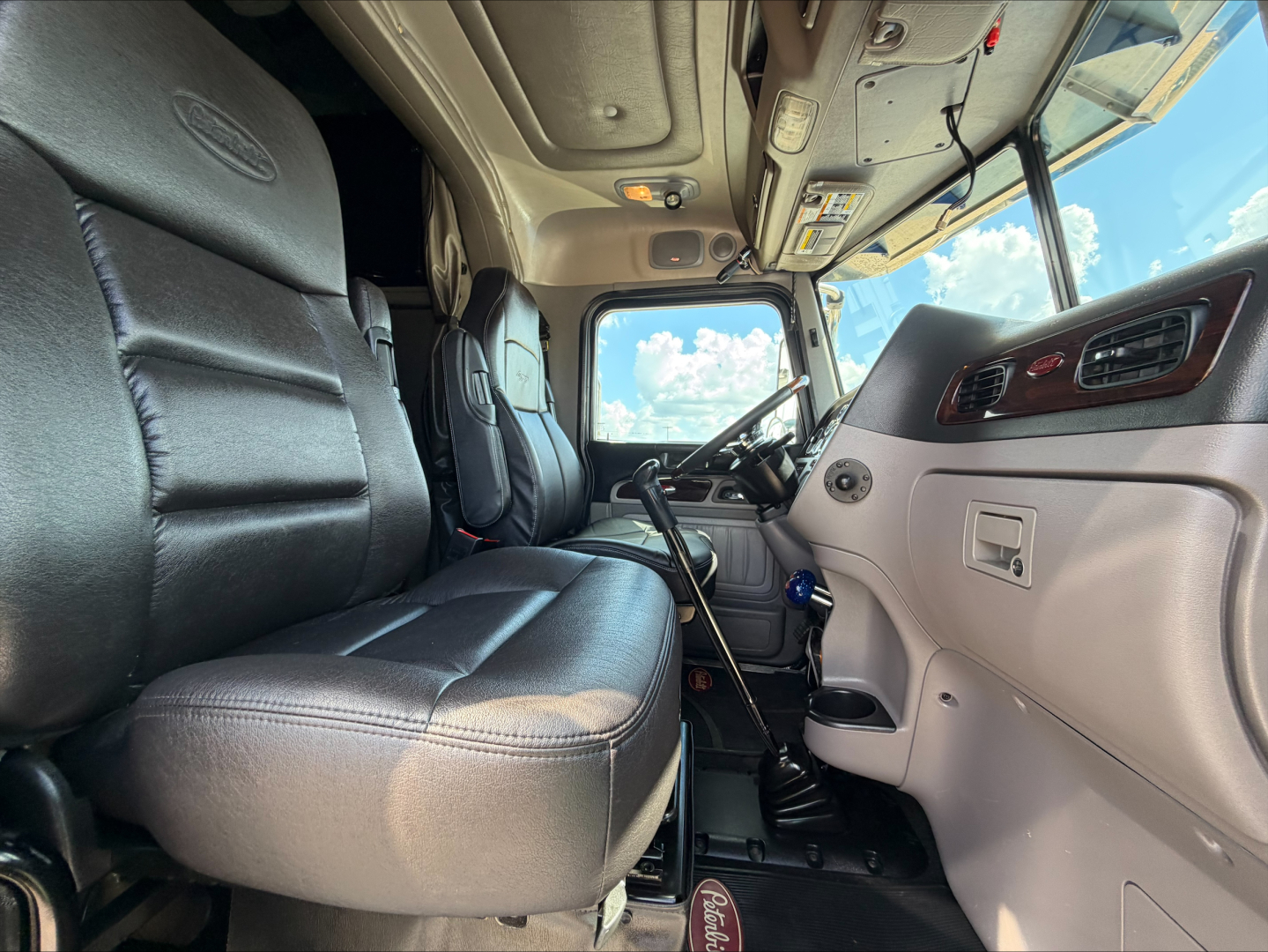 Peterbilt 389 truck interior view of driver's seat and dashboard showing steering wheel, center console, gray upholstery, and cab controls.
