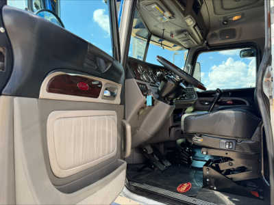 Peterbilt truck interior showing steering wheel, dash, and driver seat; door panel with light gray upholstery and wood trim; floor mat visible.