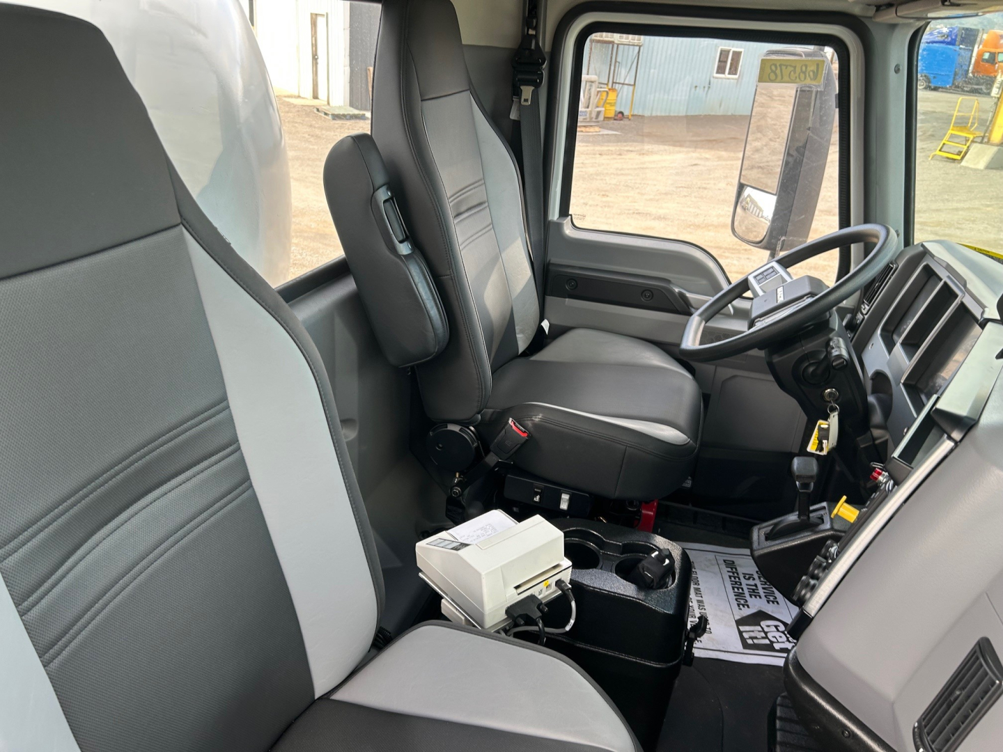 Interior of a new 2026 Mack MD7 truck cab showing driver and passenger seats, steering wheel, and dashboard with gear selector and control modules.