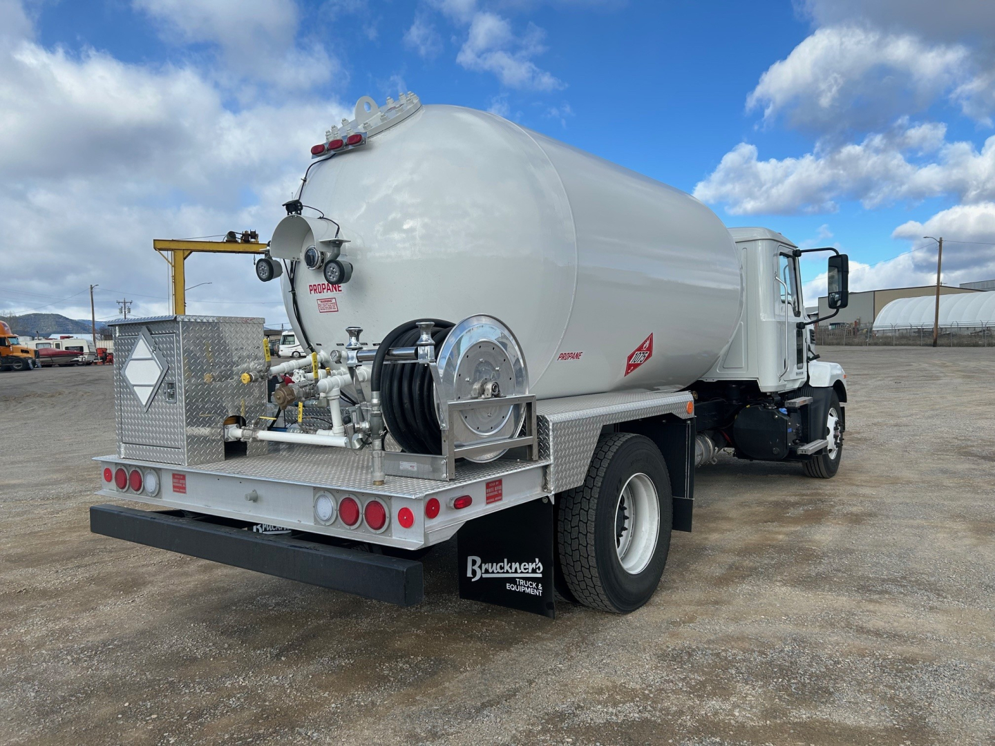Tank truck rear three-quarter view showing cylindrical stainless steel tank on a white Mack truck chassis with hose reel, safety ladders, and reflective decals.