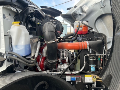 Engine bay of a 2026 Mack MD7 truck showing coolant reservoir, air intake hose, orange intercooler hose, alternator, and various hoses and labels.