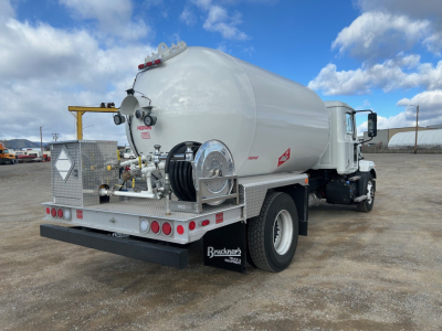 Tank truck rear three-quarter view showing cylindrical stainless steel tank on a white Mack truck chassis with hose reel, safety ladders, and reflective decals.