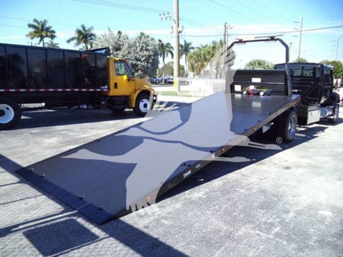 Truck deck of a rollback tow truck with a long flatbed, black metal surface, yellow tow truck in background, palm trees, overhead power lines.