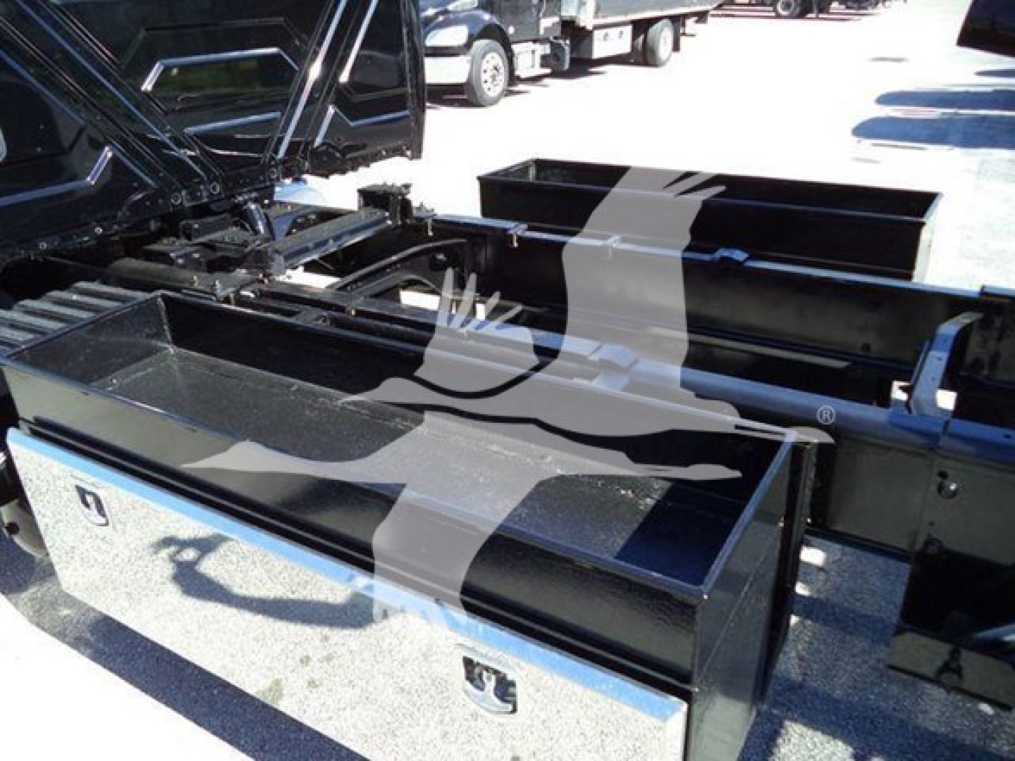 Truck rollback chassis with metal toolboxes and hydraulic components visible in the bed area. Structural frames and storage compartments under a raised hood in an outdoor lot.