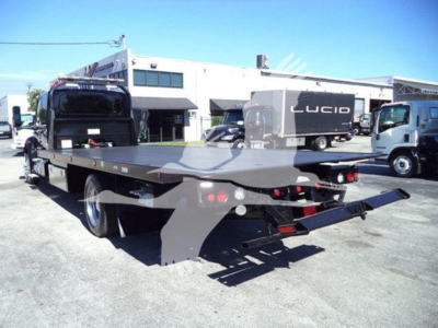 Flatbed rollback tow truck with a black hydraulic bed and rear ramps, viewed from the left-rear. Background shows dealership with other trucks and a white building.