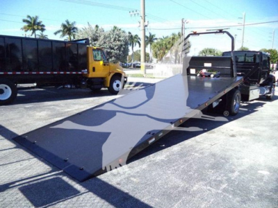 Truck deck of a rollback tow truck with a long flatbed, black metal surface, yellow tow truck in background, palm trees, overhead power lines.