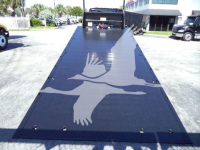 Tow truck rollback bed with bird graphic on black metal surface, viewed from front to rear, surrounding trucks in lot.
