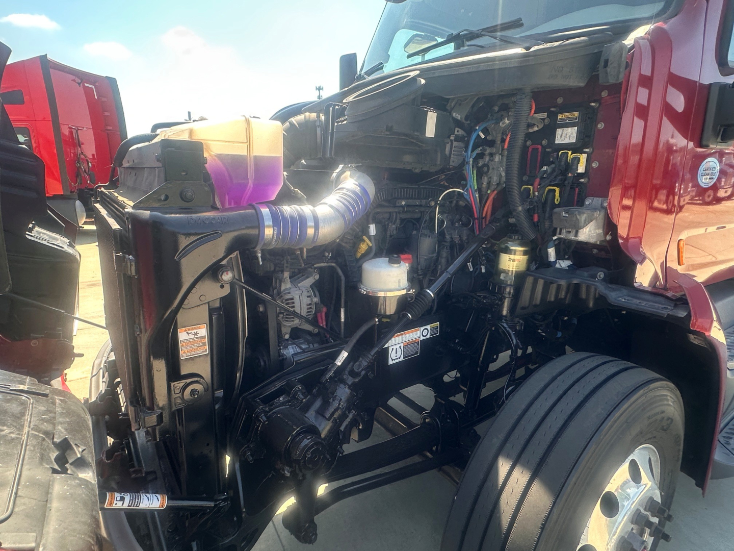 Peterbilt 579 engine bay view with exposed mechanicals, hoses, and wiring in front of the cab; components including air intake hose, coolant reservoir, and chassis visible.
