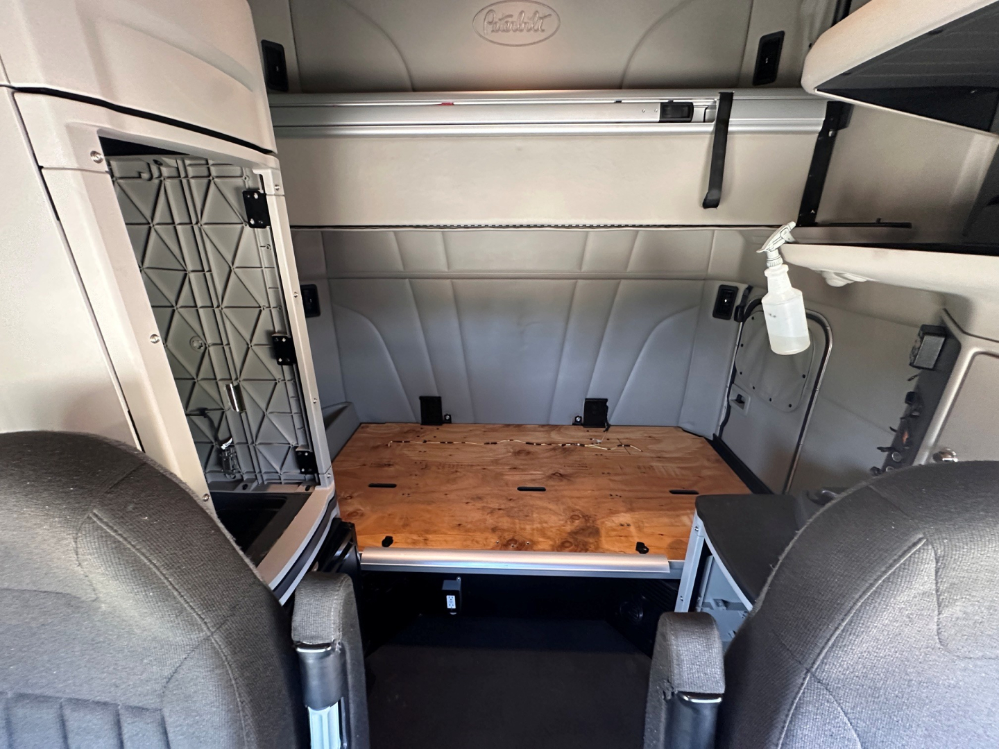 Peterbilt 579 sleeper cab interior showing cargo area with wooden floor panel, storage shelf, and spray bottle on wall; gray panels and seats in foreground.