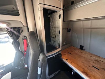 Truck cab interior with a partial sleeper and a metal partition behind the driver seat, wooden table surface on the right, and open storage compartment.