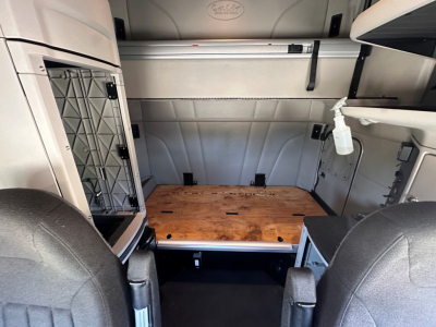 Peterbilt 579 sleeper cab interior showing cargo area with wooden floor panel, storage shelf, and spray bottle on wall; gray panels and seats in foreground.