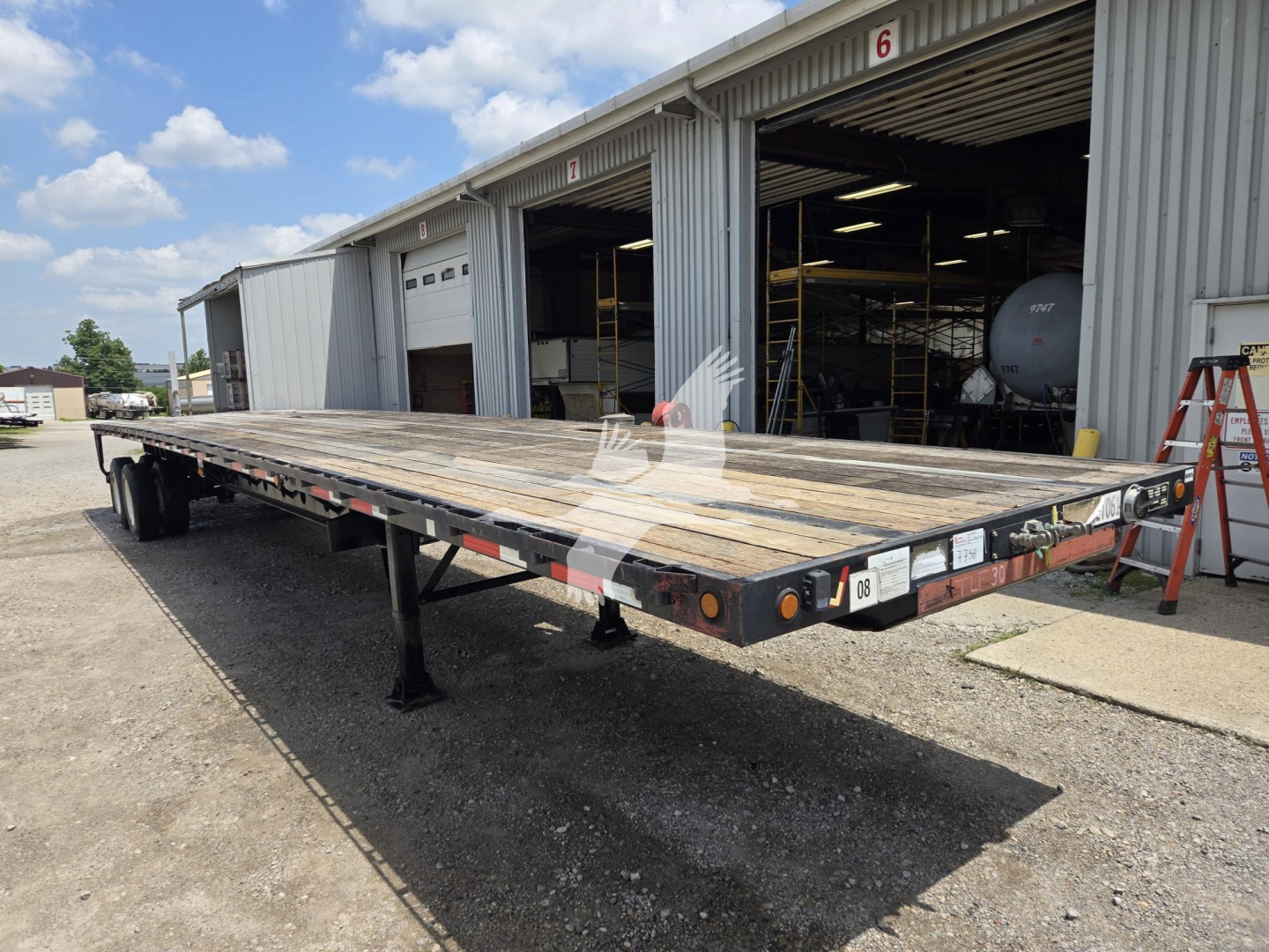Flatbed trailer view showing steel deck, dual trailing axles, and support legs in an industrial yard.