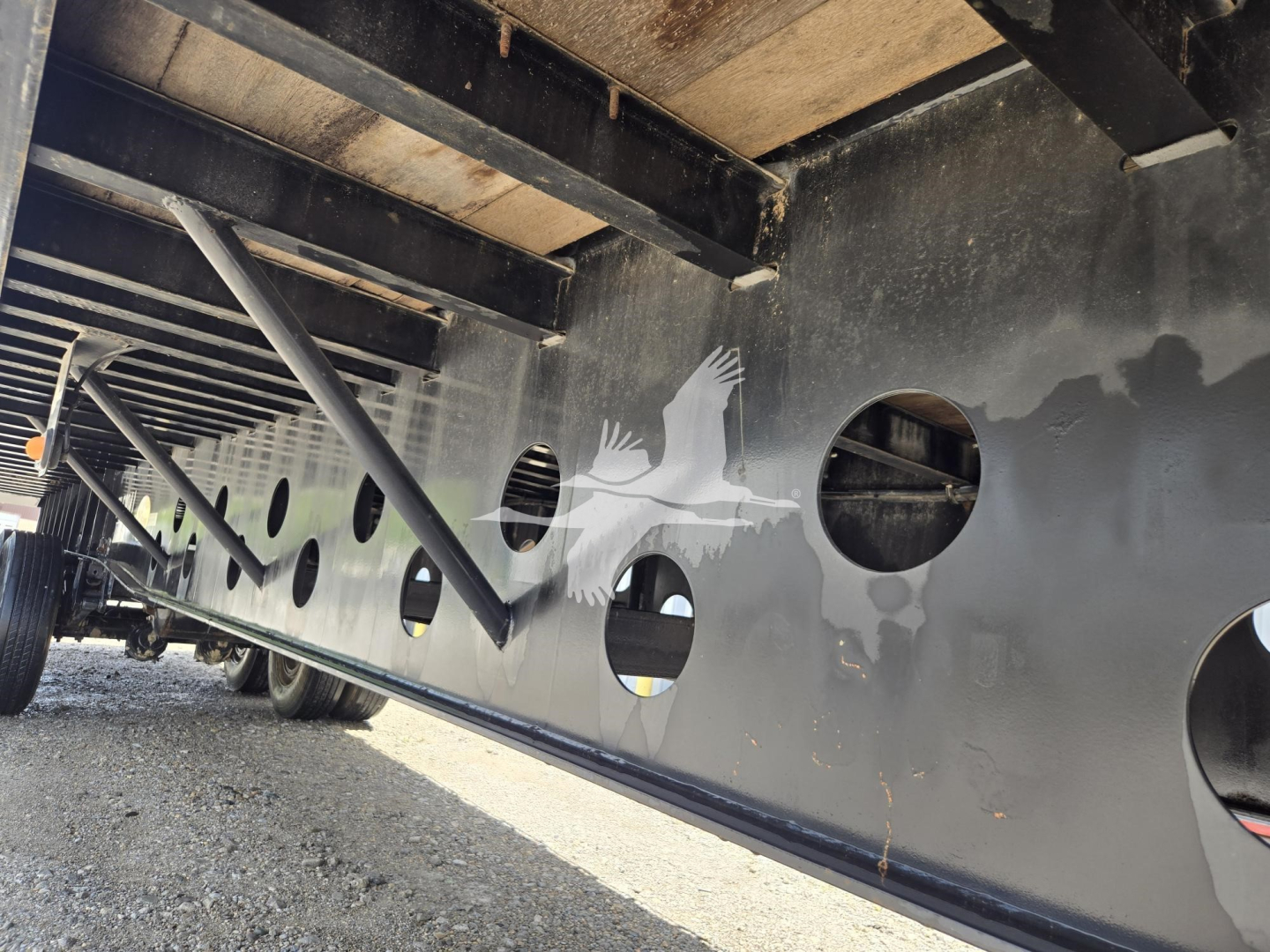 Flatbed trailer side with circular cutouts and underframe beams visible; dark metal with holes along side rail.