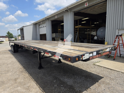 Flatbed trailer view showing steel deck, dual trailing axles, and support legs in an industrial yard.