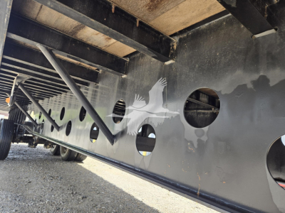 Flatbed trailer side with circular cutouts and underframe beams visible; dark metal with holes along side rail.