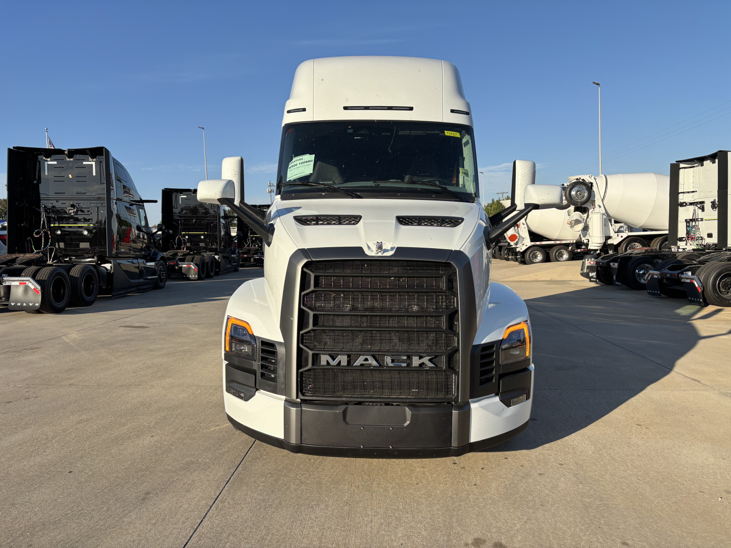 Truck front view showing Mack grille and front bumper. Hood and headlights visible; large black grille with MACK letters at center. Side mirrors and other trucks in background.