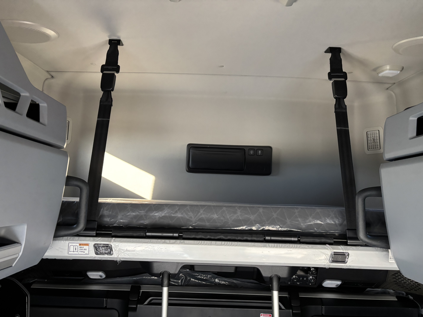 Truck cab interior showing bunk area with overhead storage and two vertical straps, side shelves, and a small center cabinet; Mack PR64T76H listing image.