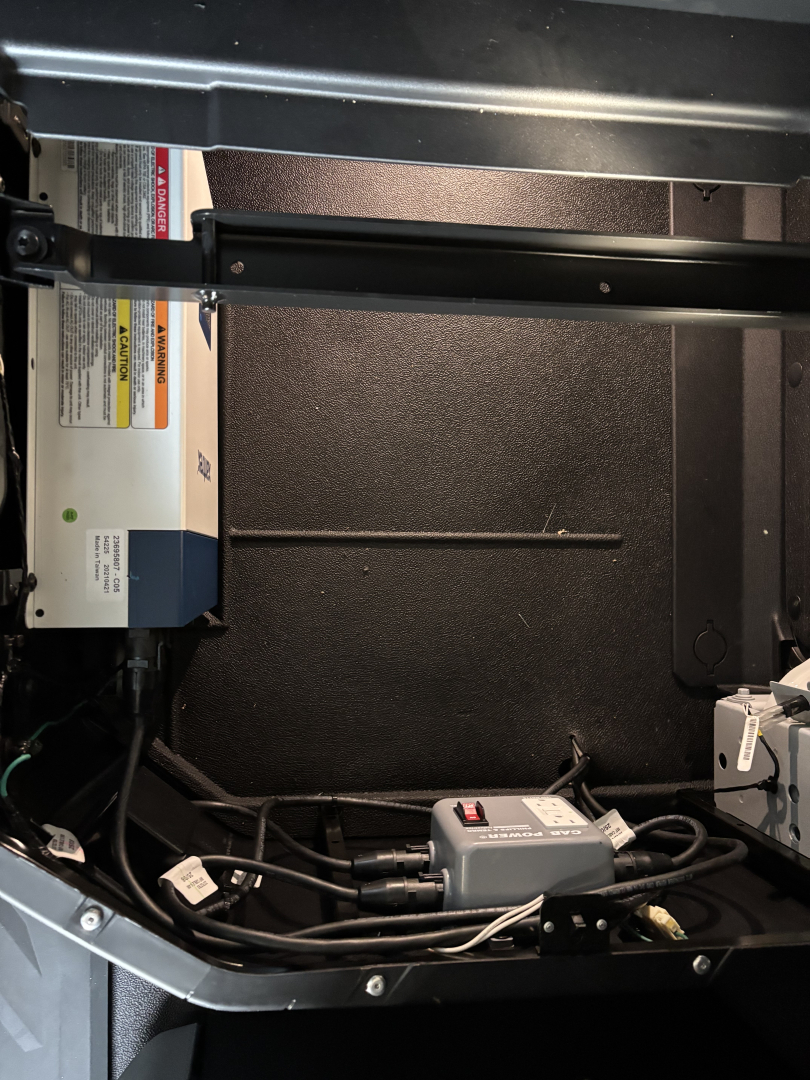 Trucks equipment compartment showing a white/blue electrical control module with warning labels, a grey control box with cables and a black interior panel.