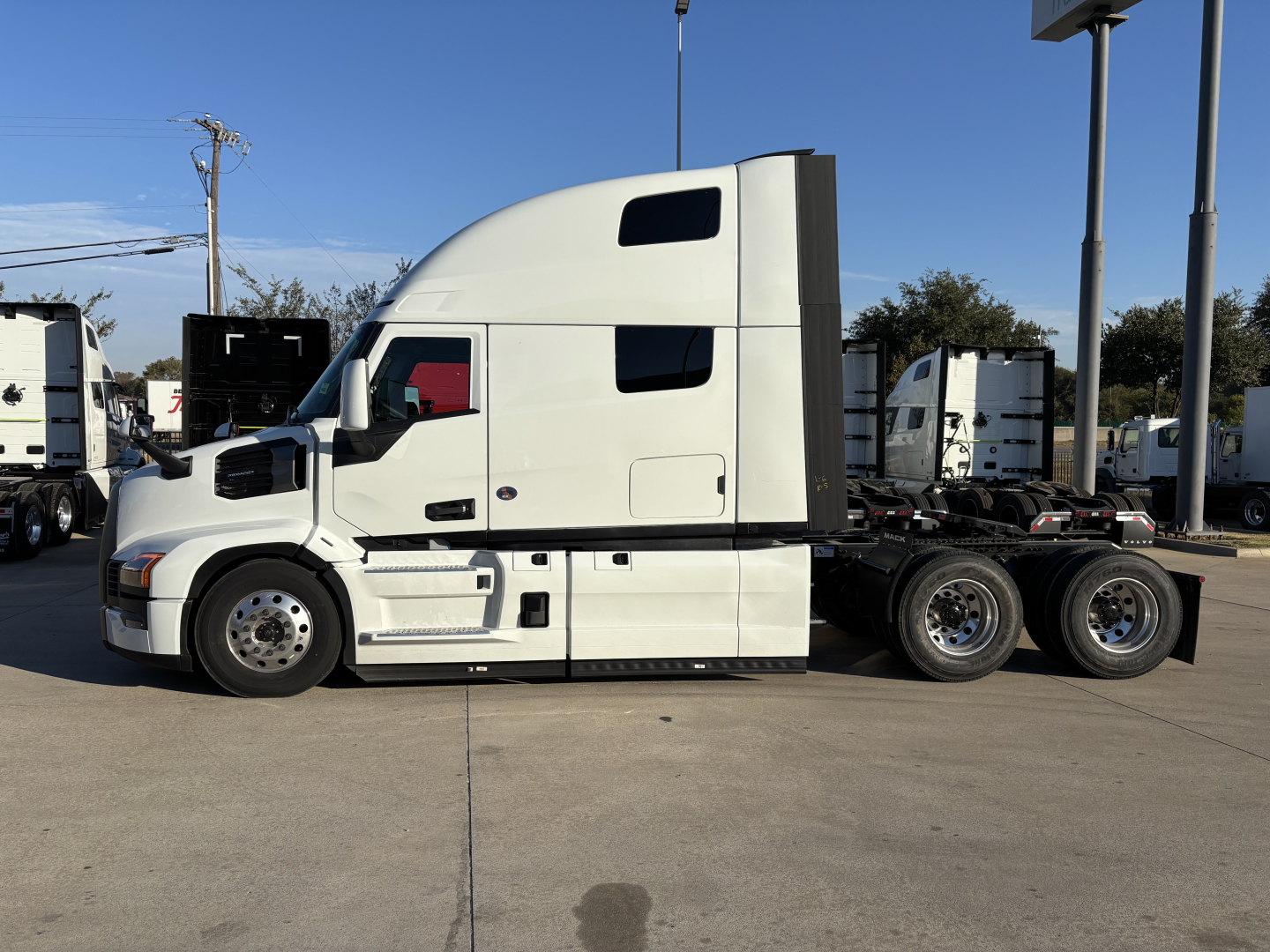 Truck side view showing a white Mack PR64T76H semi with a tall sleeper cab. No visible writing on cab; parked in lot with other trailers in background.