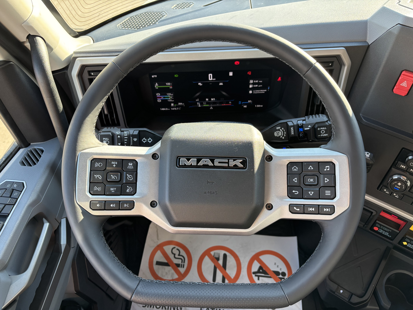 Steering wheel with Mack logo in the center, digital instrument cluster behind, control buttons on both sides of wheel. Dashboard and partial interior of a Mack PR64T76H truck cab visible.