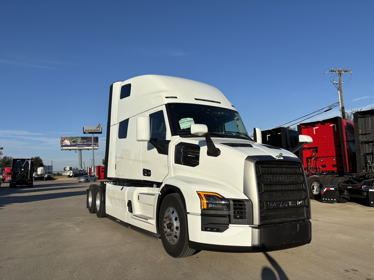 Truck in a dealership lot showing a white 2026 Mack PR64T76H conventional sleeper with aerodynamic cab, front grille, and side mirrors.