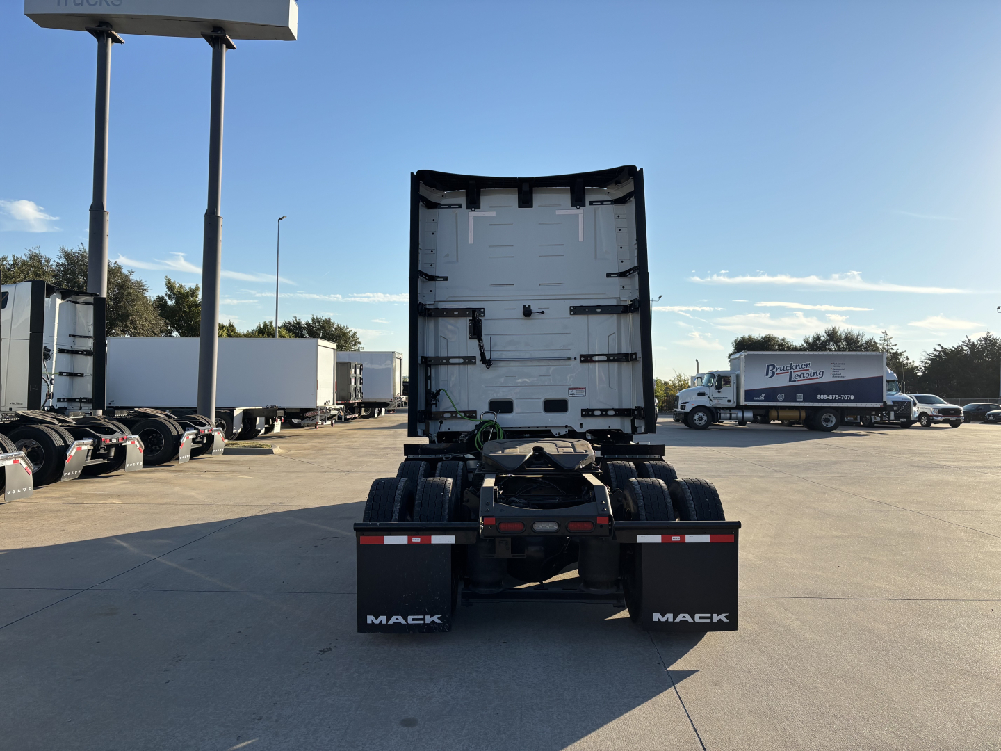 Mack PR64T76H truck tractor rear view showing fifth wheel, rear mud flaps with MACK branding, and various trailers in the lot.