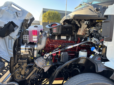 Mack PR64T76H truck engine bay showing red engine block, various hoses, reservoirs, and components.