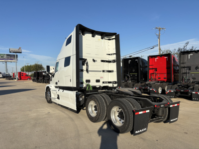 2026 Mack PR64T76H tractor truck side rear showing sleeper cab, dual rear axles, white exterior with black chassis, Mack mudflaps, and other trucks in lot.