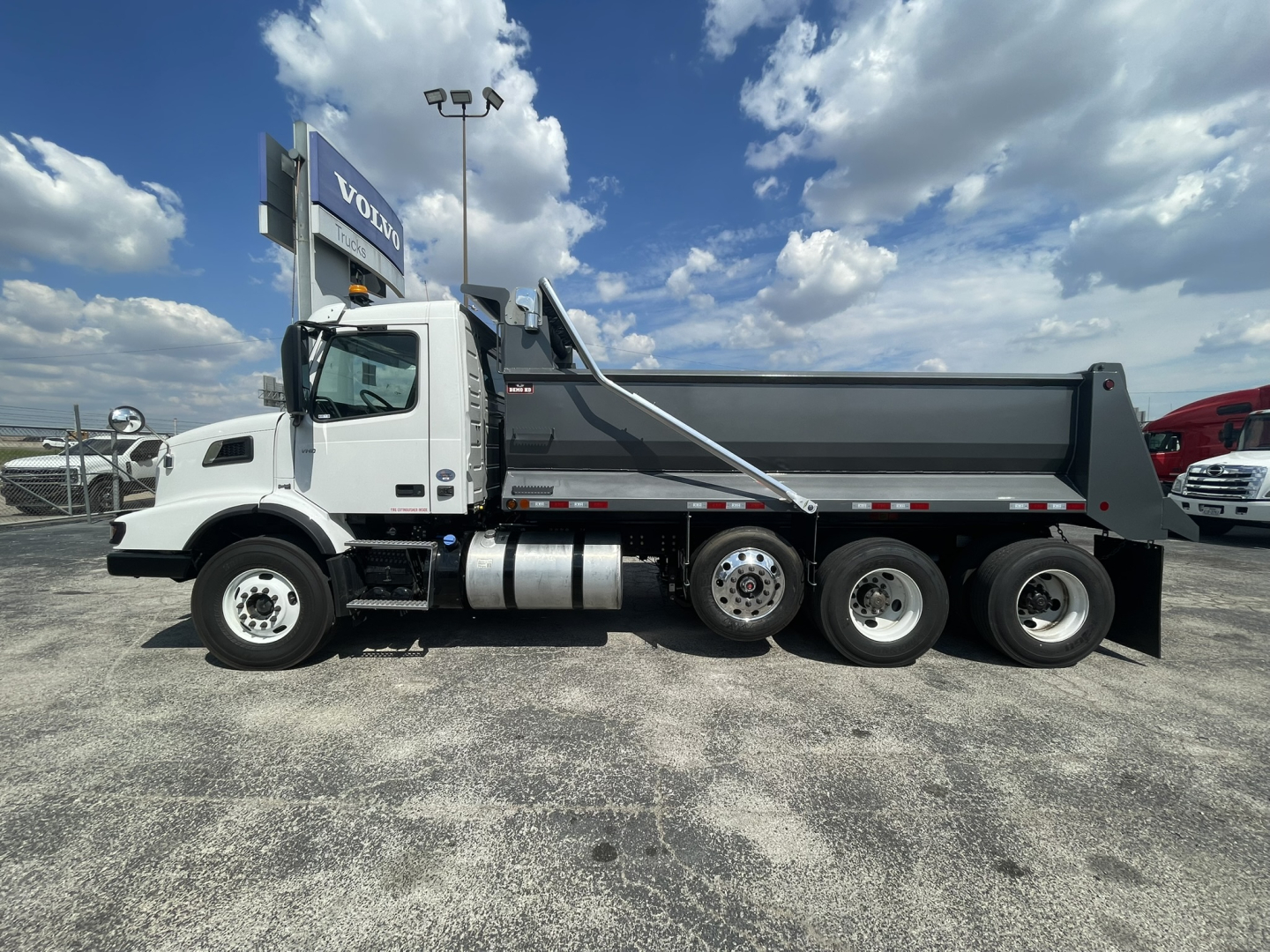 Dump truck side view showing Volvo VHD model with dumping bed, front cab, and tandem rear axles. visible texts: VOLVO on sign in background; no other legible labels on truck exterior.