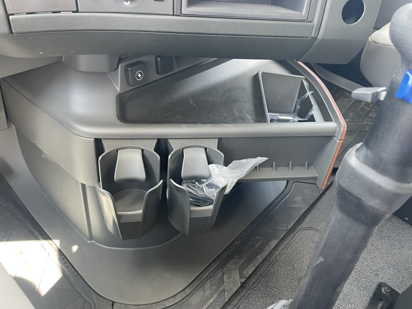 Center console area of a Volvo VHD86B300 cabin showing cup holders, small storage compartments, and a partially open lower cubby with a plastic bag inside.