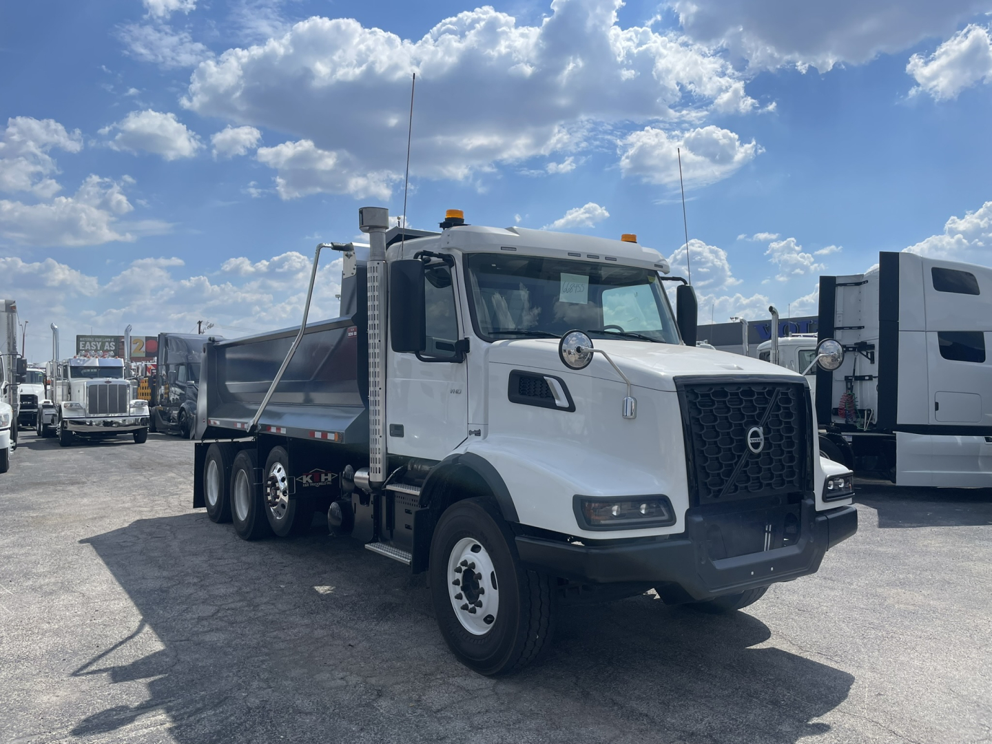 Volvo VHD86B300 dump truck in a dealership lot, white cab with black grille, chrome mirrors, and dual rear axles. Visible text includes Volvo emblem on grille and generic dealership context.